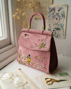 Pink handbag with floral and bird embroidery on a surface with glasses and scissors.